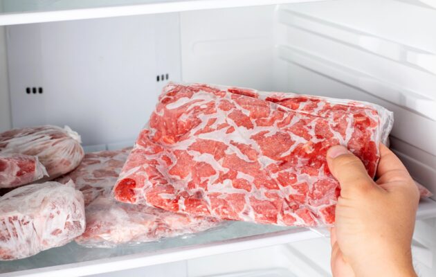 Roka izbira zamrznjeno surovo meso iz zamrzovalnika, kjer je meso shranjeno pri nizki temperaturi za ohranjanje kakovosti.